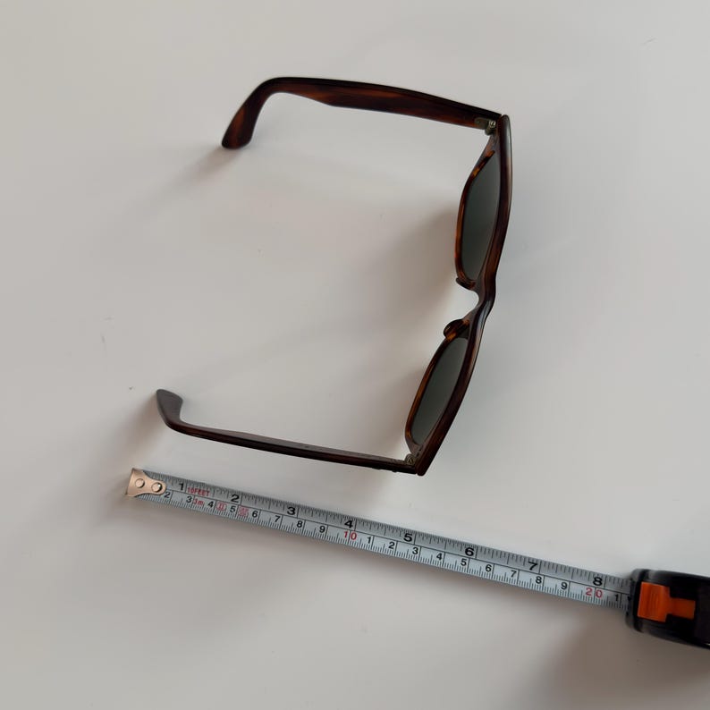 May include: Brown tortoiseshell sunglasses with dark lenses. A measuring tape is placed next to the sunglasses. The sunglasses measure approximately 6 inches in length.