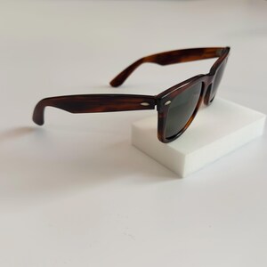 May include: Brown tortoiseshell sunglasses with dark lenses. The classic frames are set on a white surface. The sunglasses have a rectangular shape and a subtle, curved design.