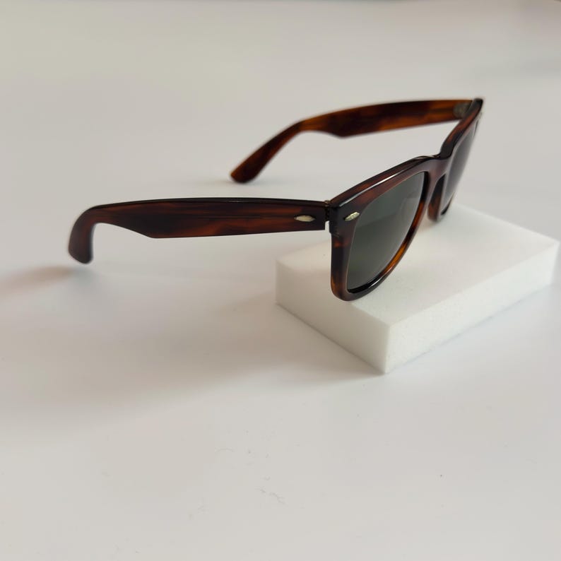 May include: Brown tortoiseshell sunglasses with dark lenses. The classic frames are set on a white surface. The sunglasses have a rectangular shape and a subtle, curved design.