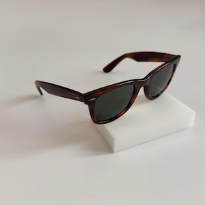 May include: Brown tortoiseshell sunglasses with dark lenses. The sunglasses have a classic rectangular frame and are resting on a white surface. The arms of the sunglasses are also brown.