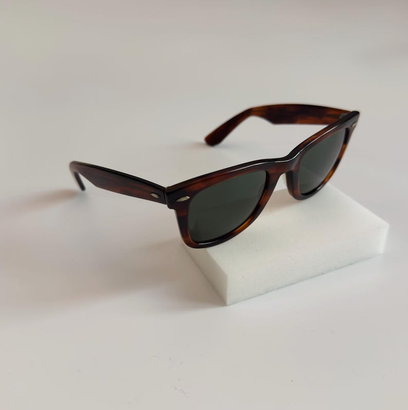 May include: Brown tortoiseshell sunglasses with dark lenses. The sunglasses have a classic rectangular frame and are resting on a white surface. The arms of the sunglasses are also brown.