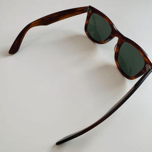 May include: Brown tortoiseshell sunglasses with green lenses. The sunglasses have a classic rectangular frame and dark brown arms. The glasses are angled, showing the shape of the frame and the lenses.
