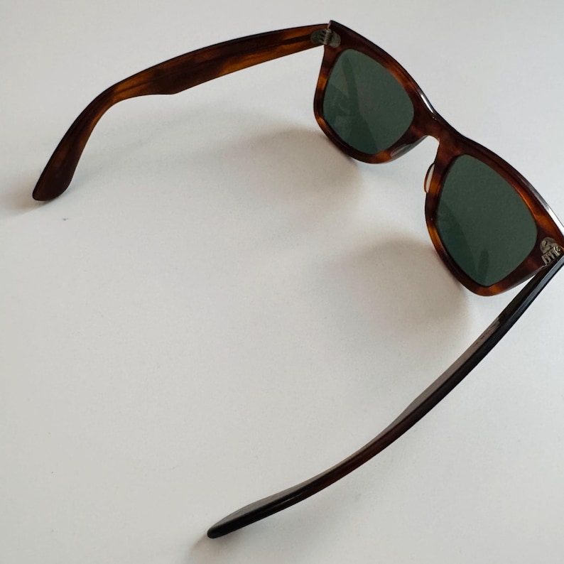 May include: Brown tortoiseshell sunglasses with green lenses. The sunglasses have a classic rectangular frame and dark brown arms. The glasses are angled, showing the shape of the frame and the lenses.