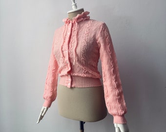 May include: A pink knitted cardigan with a button-up front and long sleeves. The cardigan has a cable knit pattern and a ruffled collar.