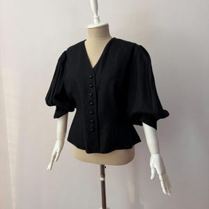 Puede incluir: Blusa negra con escote en V y cierre de botones. Presenta mangas cortas abullonadas y un bajo peplum. La prenda se exhibe en un maniquí, destacando su silueta entallada y detalles de diseño.
