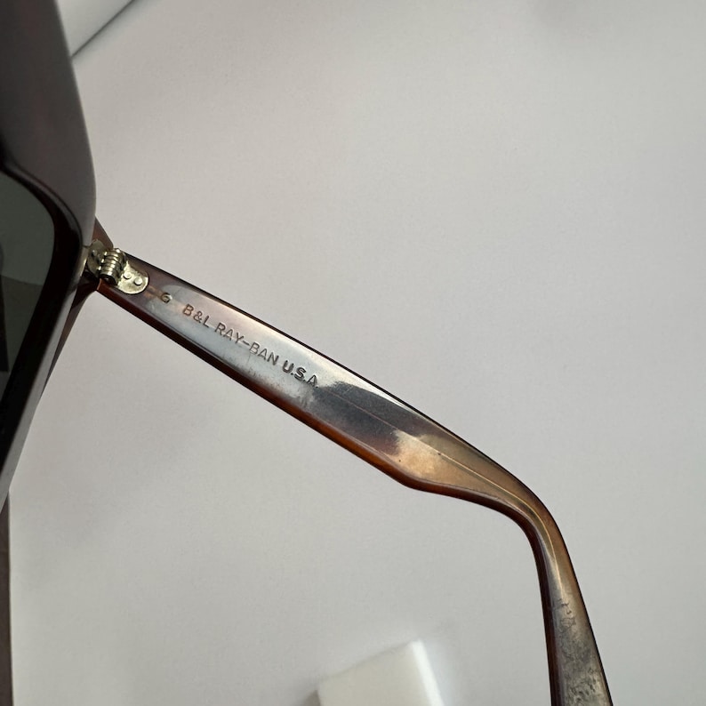 May include: Close-up of a pair of sunglasses with a brown and black frame. The text "B&L RAY-BAN U.S.A." is printed on the arm of the sunglasses. The sunglasses are set against a white background.