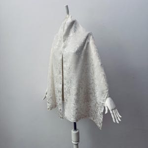 May include: A cream-colored shawl with a floral pattern is draped over a mannequin. The fabric has a subtle sheen, and the design is visible across the entire shawl. The shawl is displayed against a plain white background.