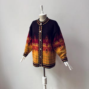 Puede incluir: Un cárdigan vintage con botones en negro, rojo y amarillo. El suéter presenta un patrón de diamantes en la parte superior y diseños florales y geométricos intrincados en las mangas y la parte inferior del cuerpo. El ribete dorado acentúa el cuello, la línea de botones y los puños.