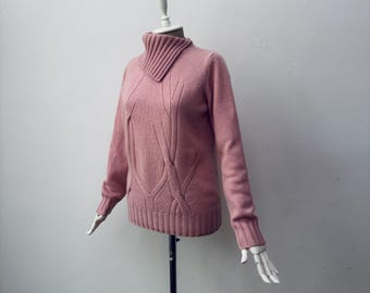 May include: A pale pink sweater with a high, ribbed collar and detailed cable knit design. The long-sleeved garment is displayed on a mannequin, highlighting its textured pattern and fitted shape. The sweater's design features vertical cable patterns.