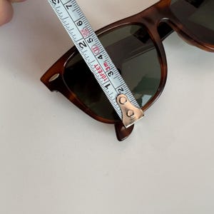 May include: Tortoiseshell sunglasses with dark lenses are measured with a tape measure. The sunglasses are approximately 5 inches in length. The frames have a classic, rectangular shape.