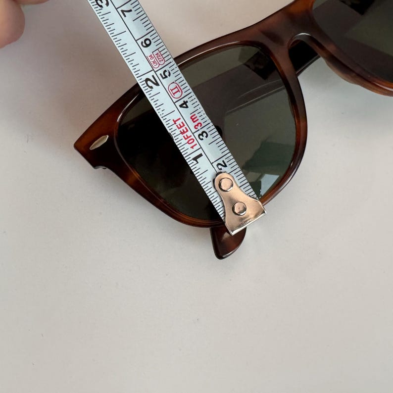 May include: Tortoiseshell sunglasses with dark lenses are measured with a tape measure. The sunglasses are approximately 5 inches in length. The frames have a classic, rectangular shape.
