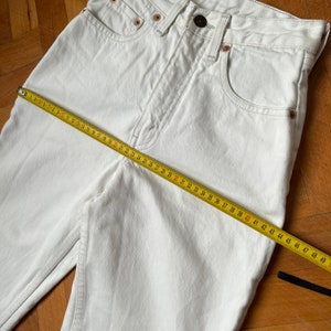 May include: Close-up of white denim jeans with a yellow measuring tape laid across the front. The jeans have a classic five-pocket design, a button closure, and copper rivets. The measuring tape shows a length of approximately 124 cm.