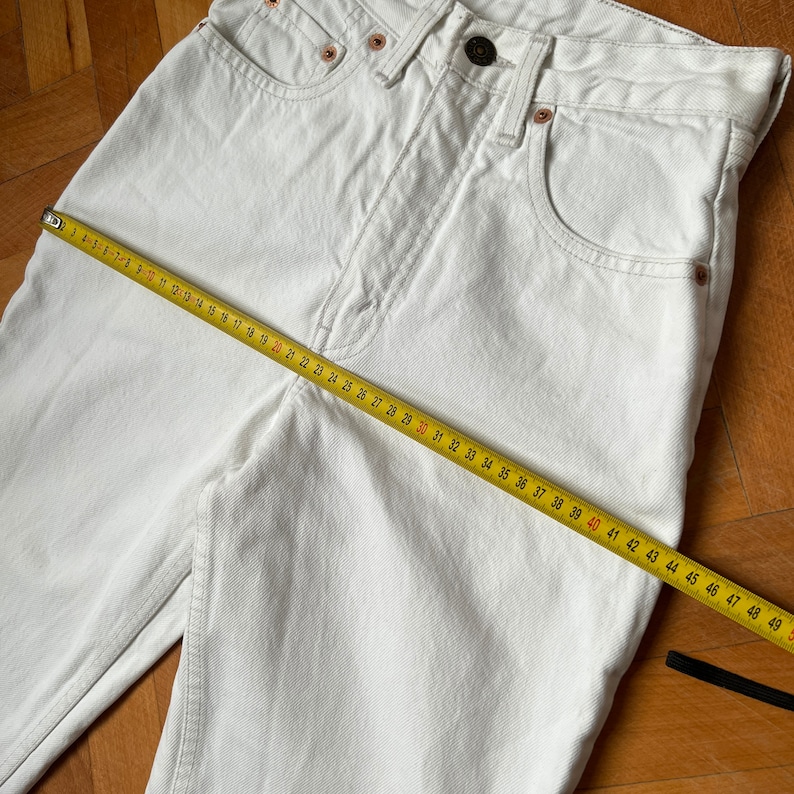 May include: Close-up of white denim jeans with a yellow measuring tape laid across the front. The jeans have a classic five-pocket design, a button closure, and copper rivets. The measuring tape shows a length of approximately 124 cm.