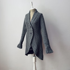 Puede incluir: Un cárdigan gris de manga larga con cuello chal y cierre de botones. El suéter presenta detalles de punto trenzado y acanalado, con puños acampanados. La prenda se exhibe en un maniquí sobre un fondo blanco liso.