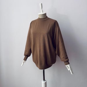 Puede incluir: Un suéter marrón de manga larga con cuello alto se muestra en un maniquí. El suéter tiene un corte holgado con puños y dobladillo acanalados. El maniquí tiene cabeza y brazos blancos y está montado sobre un soporte metálico sobre un fondo blanco.