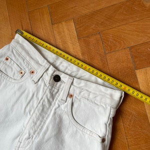 May include: Close-up of white denim jeans with a button and copper rivets. A yellow measuring tape is laid across the waistband, showing a measurement of approximately 89 cm.