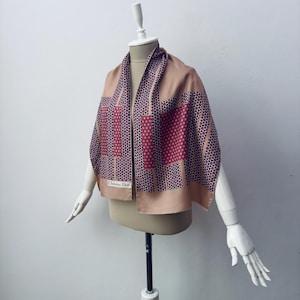 Puede incluir: Un pañuelo de seda color beige con un estampado geométrico en burdeos, negro y blanco. El pañuelo está drapeado sobre un maniquí y lleva la etiqueta "Christian Dior".