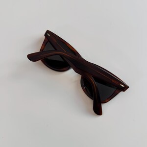 May include: Brown tortoiseshell sunglasses with dark lenses. The sunglasses have a classic frame design with a slightly angled browline and thick temples. The arms are curved for a comfortable fit. The sunglasses are set against a white background.