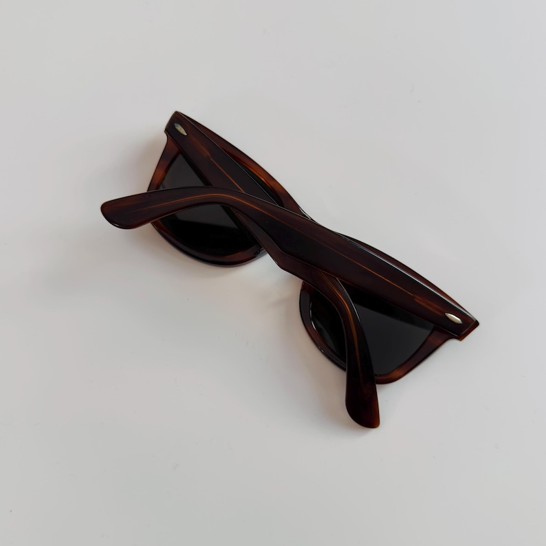 May include: Brown tortoiseshell sunglasses with dark lenses. The sunglasses have a classic frame design with a slightly angled browline and thick temples. The arms are curved for a comfortable fit. The sunglasses are set against a white background.