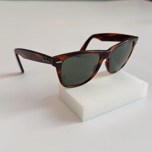 May include: Classic tortoiseshell sunglasses with dark green lenses. The sunglasses have a rectangular frame and are displayed on a white block. The Ray-Ban logo is visible on the temple.