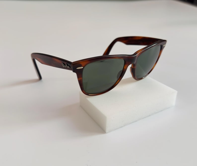 May include: Classic tortoiseshell sunglasses with dark green lenses. The sunglasses have a rectangular frame and are displayed on a white block. The Ray-Ban logo is visible on the temple.