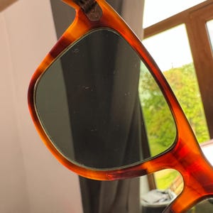 May include: Close-up of sunglasses with a tortoiseshell pattern frame. The lenses are dark, and the frame has a gradient of brown and orange hues. The sunglasses are held up, with a blurred background of a window and greenery.