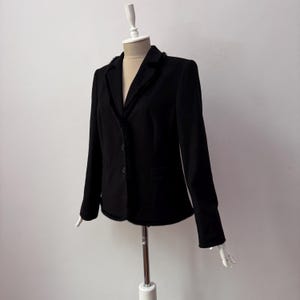 Może przedstawiać: Czarny blazer ze lekko strzępionym kołnierzem i dwoma guzikami. Blazer ma jedną kieszeń po lewej stronie.