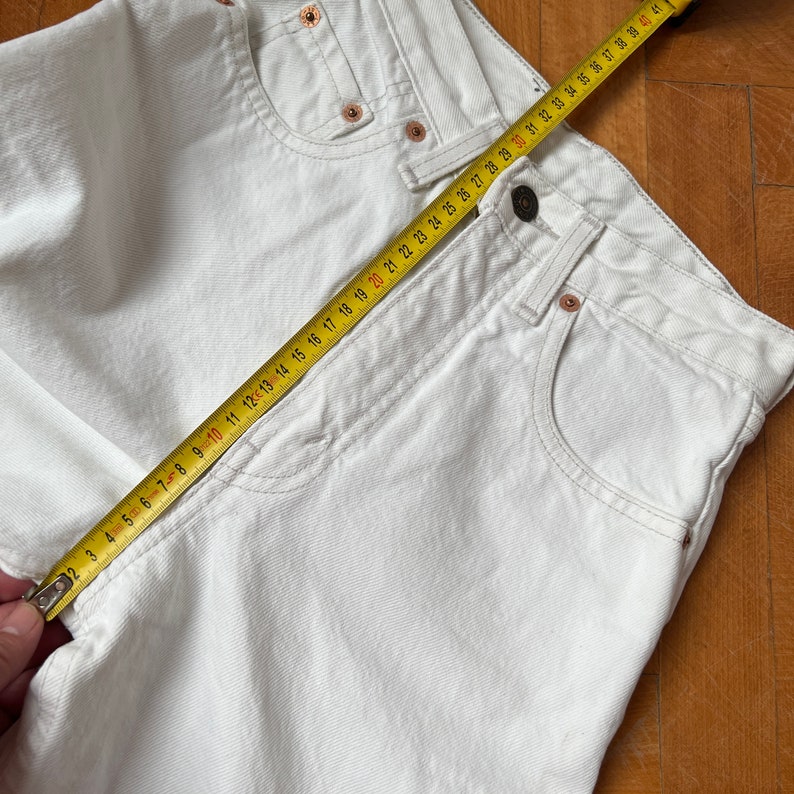May include: Cream-coloured denim jeans with a classic five-pocket design. A yellow measuring tape is laid across the jeans, showing measurements in centimetres. The jeans have copper rivets and a button closure.