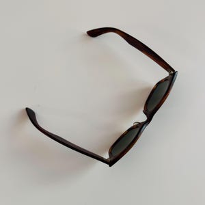 May include: Sunglasses with a tortoiseshell pattern frame and dark lenses. The arms of the sunglasses are a dark brown color. The sunglasses are angled, showing the shape of the lenses and the frame.