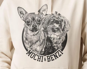以下が含まれることがあります： 胸元に白黒の円形グラフィックが施されたクリーム色のフード付きスウェットシャツ。グラフィックには2匹の犬が描かれており、円形デザインの下端に太字のサンセリフ体で「MOCHI & BENZI」という文字がプリントされています。