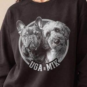 May include: Black sweatshirt featuring a circular graphic of two dogs, a French Bulldog and a terrier mix. The graphic is in grayscale with the words "UGA & MIK" in white. The sweatshirt is worn by a person.