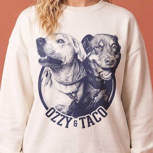 May include: A cream-colored crewneck sweatshirt featuring a circular graphic print of two dogs with the text OZZY & TACO printed in dark blue below the illustration. The sweatshirt has long sleeves with ribbed cuffs and a ribbed neckline.