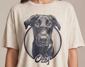 May include: Beige t-shirt featuring a black and white illustration of a dog within a circular frame. The dog's name, "Ozzy," is written in a cursive font below the image. The shirt has short sleeves and a relaxed fit.