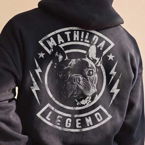 May include: A black hooded sweatshirt featuring a white graphic on the back. The design includes a French Bulldog portrait inside a circular frame with the text MATHILDA at the top and LEGEND at the bottom, flanked by two lightning bolt symbols.