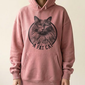 Puede incluir: Una sudadera con capucha de color rosa polvoriento que presenta un gr&aacute;fico circular negro en el pecho. El gr&aacute;fico muestra un gato de pelo largo dentro de un c&iacute;rculo con el texto A FAT CAT impreso en una fuente negrita sin remates.