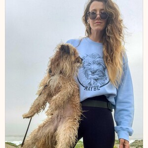 May include: A light blue crewneck sweatshirt featuring a black circular graphic of a scruffy dog's face with the word MAYBE printed in black capital letters underneath the dog's chin. The sweatshirt has long sleeves and a ribbed collar and cuffs.