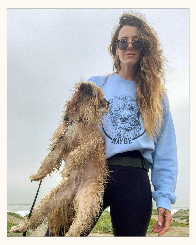 May include: A light blue crewneck sweatshirt featuring a black circular graphic of a scruffy dog's face with the word MAYBE printed in black capital letters underneath the dog's chin. The sweatshirt has long sleeves and a ribbed collar and cuffs.