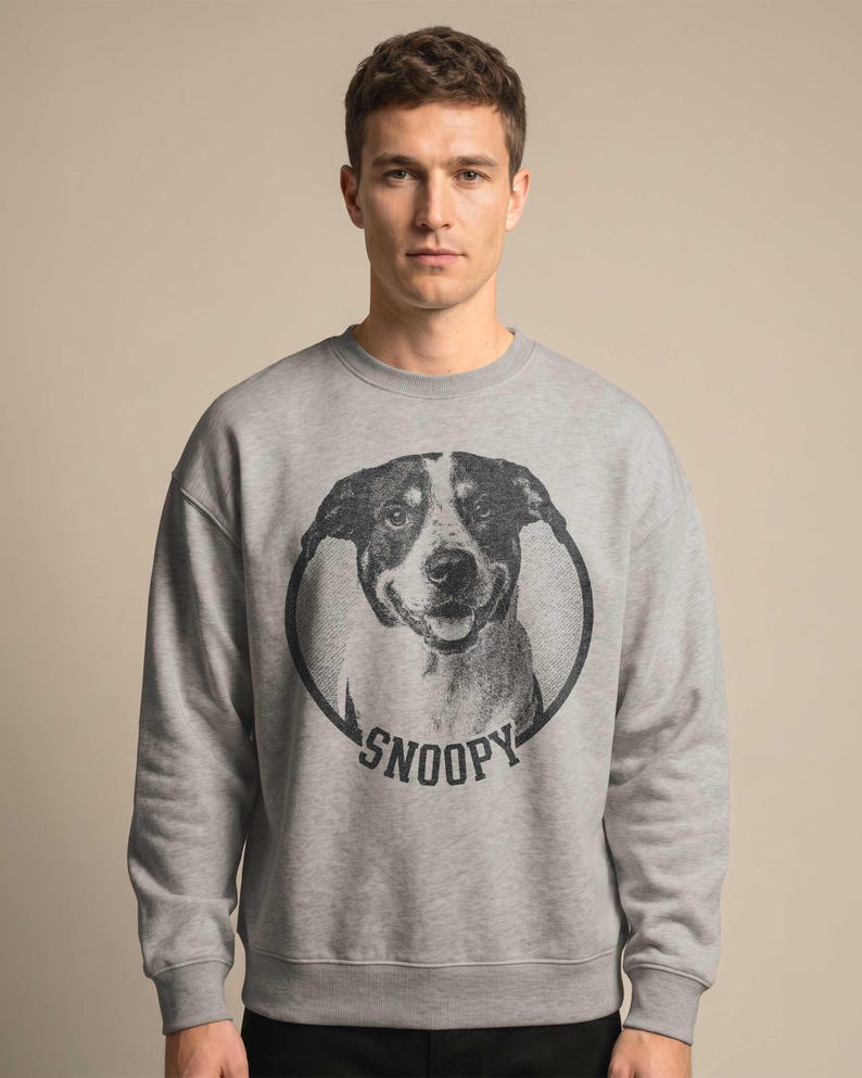 May include: A gray sweatshirt featuring a black and white dog portrait inside a circular frame. The word "SNOOPY" is printed in black below the dog image. The sweatshirt is a casual style.