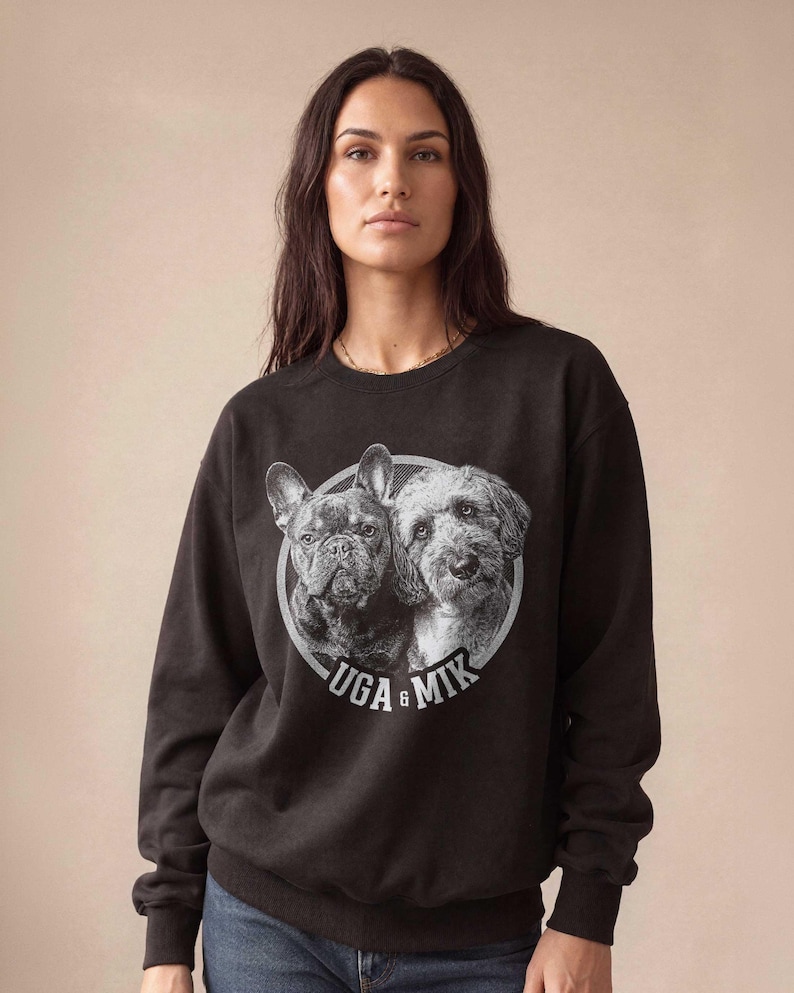 May include: Black sweatshirt featuring a circular graphic of two dogs, a French Bulldog and a terrier mix. The graphic is in grayscale with the words "UGA & MIK" in white. The sweatshirt is worn by a person.