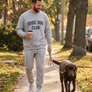 May include: A light gray crewneck sweatshirt featuring the dark blue text GOOD DOG CLUB on the chest, paired with matching light gray sweatpants and white athletic sneakers. The outfit is worn while holding a coffee cup and a dog leash.