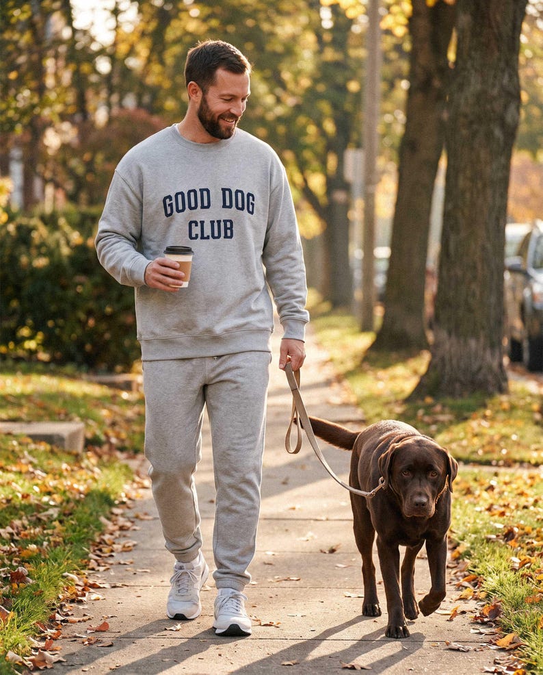 May include: A light gray crewneck sweatshirt featuring the dark blue text GOOD DOG CLUB on the chest, paired with matching light gray sweatpants and white athletic sneakers. The outfit is worn while holding a coffee cup and a dog leash.