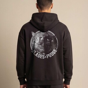 Puede incluir: Una sudadera con capucha negra que presenta un gr&aacute;fico circular en la parte posterior. El gr&aacute;fico muestra una ilustraci&oacute;n en escala de grises de un gato atigrado y un gato negro juntos, con el texto "BLADES & PUDGE" en letras blancas.