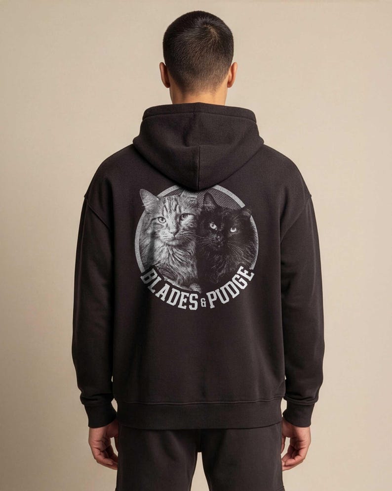 Puede incluir: Una sudadera con capucha negra que presenta un gr&aacute;fico circular en la parte posterior. El gr&aacute;fico muestra una ilustraci&oacute;n en escala de grises de un gato atigrado y un gato negro juntos, con el texto "BLADES & PUDGE" en letras blancas.
