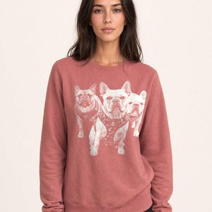 May include: A dusty rose crewneck sweatshirt featuring a white graphic print of three French Bulldogs on the front. The dogs are depicted in a standing position, with the central dog wearing a harness. The sweatshirt has long sleeves and ribbed cuffs.