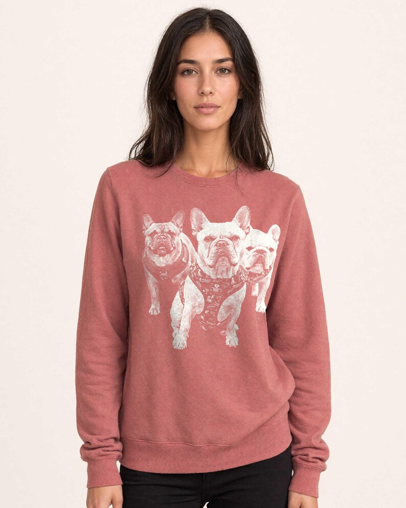 May include: A dusty rose crewneck sweatshirt featuring a white graphic print of three French Bulldogs on the front. The dogs are depicted in a standing position, with the central dog wearing a harness. The sweatshirt has long sleeves and ribbed cuffs.