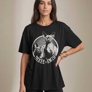 Puede incluir: Camiseta negra con un gr&aacute;fico de dos gatos Sphynx dentro de un c&iacute;rculo blanco. El texto "Dixee & Enzo" est&aacute; impreso alrededor del c&iacute;rculo. La camiseta tiene mangas cortas y un corte holgado.