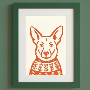 Op de afbeelding: Een ingelijste print met een gestileerde oranje illustratie van een hond die een trui met een wit bloemenpatroon draagt. De hond heeft puntige oren en staat centraal op een cr&egrave;mekleurige achtergrond. Het kunstwerk zit in een donkergroene lijst.