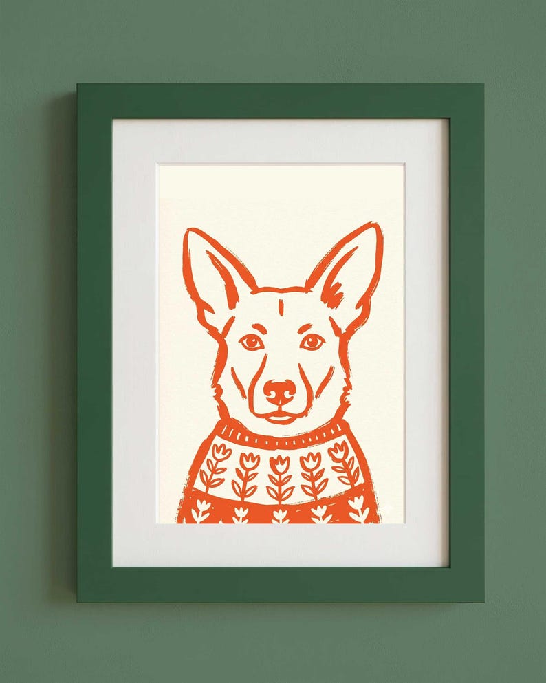 Op de afbeelding: Een ingelijste print met een gestileerde oranje illustratie van een hond die een trui met een wit bloemenpatroon draagt. De hond heeft puntige oren en staat centraal op een cr&egrave;mekleurige achtergrond. Het kunstwerk zit in een donkergroene lijst.