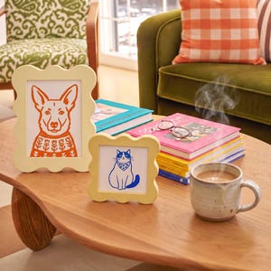 Op de afbeelding: Twee lichtgele fotolijsten met een geschulpte rand. De grotere rechthoekige lijst bevat een oranje illustratie van een hond in een trui met patroon. De kleinere vierkante lijst bevat een blauwe illustratie van een kat met een bandana.
