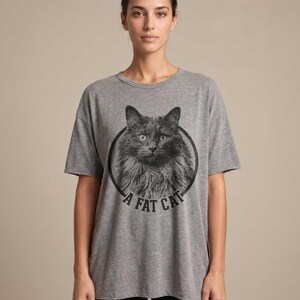 Puede incluir: Camiseta gris con una ilustraci&oacute;n en blanco y negro de un gato peludo dentro de un c&iacute;rculo negro. El texto "A FAT CAT" est&aacute; impreso debajo de la imagen del gato. La camiseta tiene mangas cortas y un corte holgado.